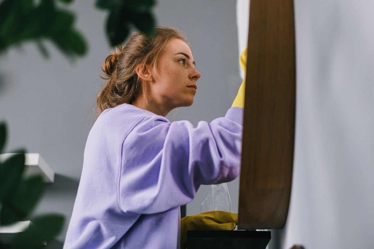 services-04 Vue de côté d’une jeune femme concentrée, portant des gants en latex, debout et nettoyant un miroir dans une pièce lumineuse en journée