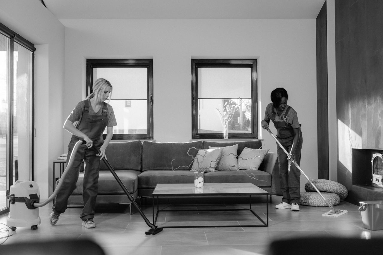why-choose-us Deux femmes en tenue de travail nettoient un salon moderne à l’aide d’un aspirateur et d’une serpillière.